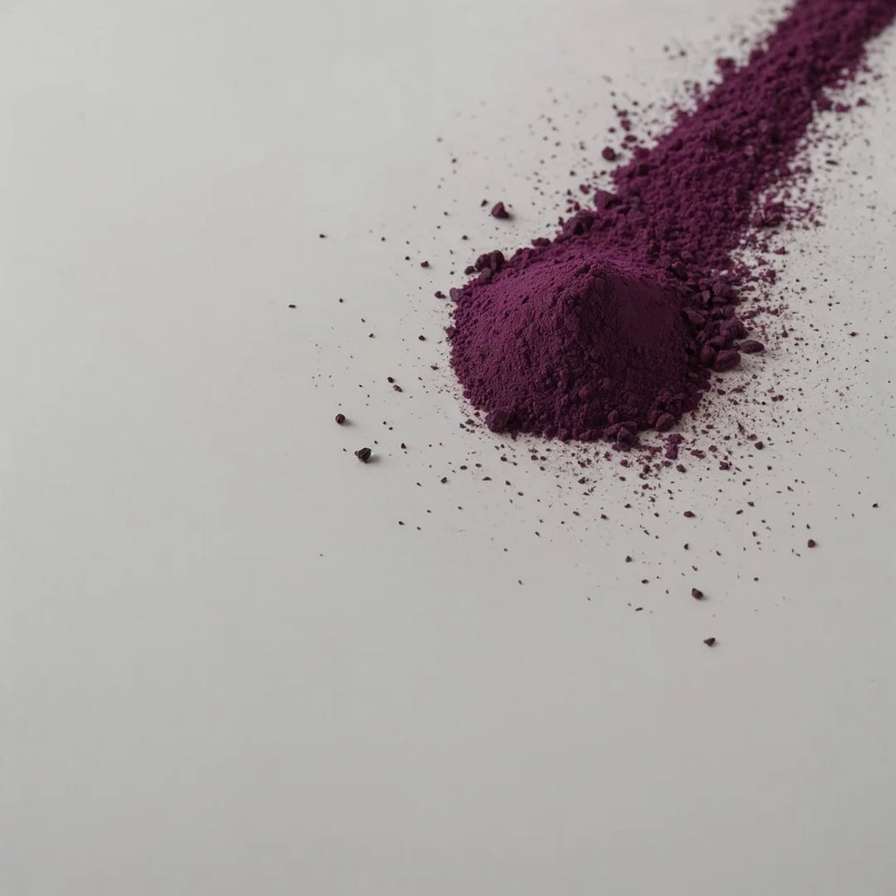 Monticule de poudre de roselle violette avec des particules éparpillées sur une surface blanche, illustrant sa légèreté et sa texture raffinée.